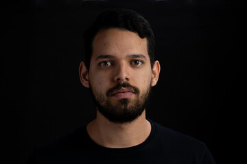 Young man frontal head shot with black background. He is serious. He has a casual look with beard...