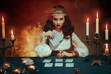 Fantasy beautiful magnificent girl in image of a gypsy sits at table in dark gothic room. Red costume, vintage clothing, art black make up. Fortune teller woman reading future on magical tarot cards. - obrazy, fototapety, plakaty