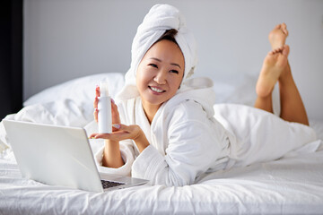 woman lying on comfortable bed at home, using cosmetics, showing bottle to someone via webcam, look at camera smiling, in bathrobe and towel