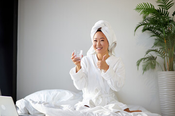 korean woman in abthrobe and towel sits on bed with glass of fresh water, enjoy. female begins the...