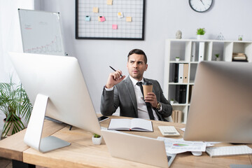 thoughtful trader holding coffee to go and pen near notebook and monitors at workplace