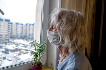 Lonely old woman in a medical mask looks out the window during the lockdown. A depressed lady at home during the covid-19 pandemic.