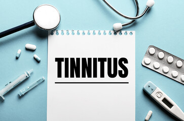 The word TINNITUS written on a white notepad on a blue background near a stethoscope, syringe, electronic thermometer and pills. Medical concept