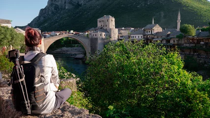 Cercles muraux Stari Most A woman with a backpack and a tripod on her back is sitting on a stone wall and looking at the Old bridge (Stari most) in Mostar, Bosnia and Herzegovina.