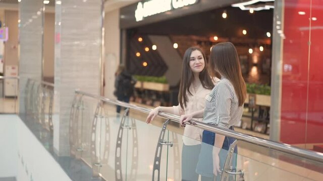 two young women are talking by the glass railing in the shopping center.