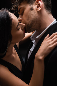 elegant woman with closed eyes kissing with man in suit isolated on black
