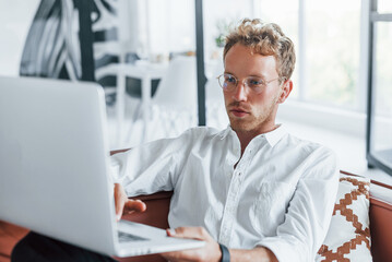 With laptop. Caucasian young guy in elegant white shirt indoors at home