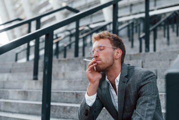 Sits and smokes cigarette. Young successful businessman in grey formal wear is outdoors in the city