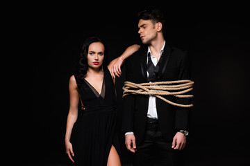 brunette woman in dress standing with tied submissive man isolated on black