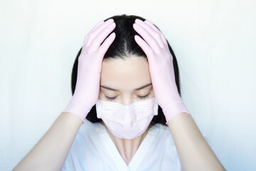 A woman in a protective mask and gloves on a white background holds her head with two hands. The stress due to the coronavirus.