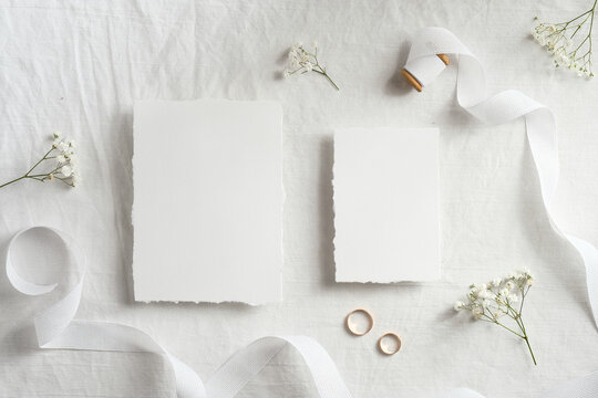 Blank wedding invitations cards mockups with space for text, golden rings, ribbon flowers on white textile background. Flat lay, top view.