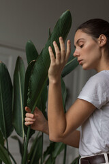 Young woman gardener touching lush houseplant. Woman is wearing white trousers and a T-shirt. Indoor cozy garden. Love plants, hobby, home gardening concept, freelance, small business.