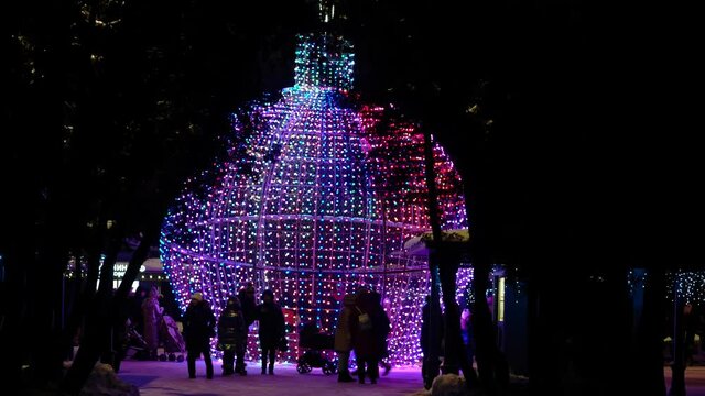 people have fun and take pictures around the festive illumination in the form of a Christmas tree toy in a night park in winter
