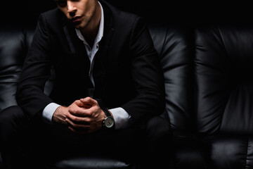 cropped view of confident man in suit sitting on black leather sofa