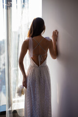 Portrait of gorgeous happy luxury stylish brunette bride in silk robe, standing at the window in skycraper. Beautiful details on wedding dress.  © Dragan