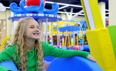 A young blonde girl of European appearance with blond curly hair smiles at the children's playground and entertainment center.