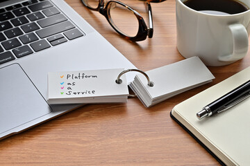 There's a laptop computer, a notebook, a pen and an open vocabulary book on the desk. The word Platform as a Service is written on it.