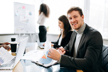 Portrait of smiling male manager in formal wear at briefing meeting. Successful business man learns financial graphs, documents. Caucasian male employee looks directly at the camera and smile