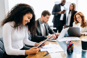 Successful african american lady at business meeting. Beautiful female employer, learning financial graphs, doing paperwork, smile. Young adult manager sitting at the desk in modern office