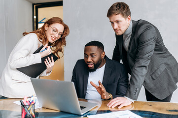 Happy multiracial business people in office using a laptop, look at the screen. Successful coworkers reading good news, get agreement for a project. Young adult employees working together, smiling