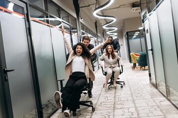 Young adult colleagues take a break from work. Friendly coworkers make a race on office chairs....