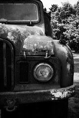 Fototapeta premium Black and white detail of a vintage pick up truck showing head light and a section of grill. Truck has holes caused by rust. It has peeling, faded and scratched paint. The head light has broken glass