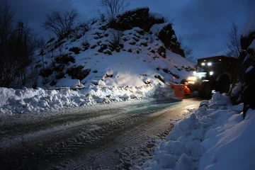 Fotobehang snowplow passes through the snowy mountain streets in the evening to clean up the road traffic © Alessia