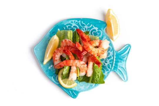 Overhead View Of Large Cooked Peeled Tiger Shrimps On A Blue Fish  Shaped Plate With Lemon Wedges And Tangy Sauce Isolated On White