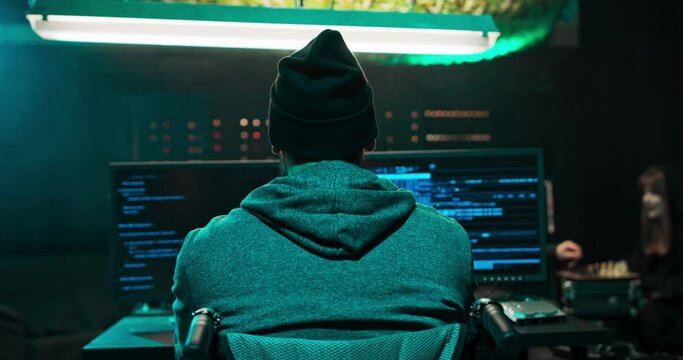 Shot From The Back To Man In A Wheelchair, A Hooded Hacker Works On A Computer With Data Displayed On Screens In A Secret Hideout. In The Background You Can See Monitors And Temporary Servers.