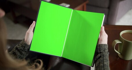 Over the shoulder slider shot of a young woman reading a book with green screen pages and a cup of coffee - Powered by Adobe