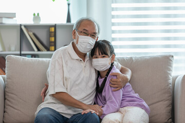 Asian Elderly grandfather hug grandchild and wearing face mask on sofa at home protect virus...
