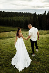 Stylish couple are walking at the field. Handsome groom holding hand of gorgeous bride, enjoy togetherness. Beautiful woman with attractive man on wedding day, posing, photoshoot concept