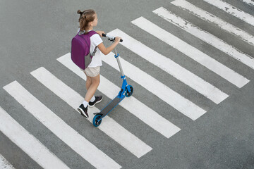 Schoolgirl carrying scooter and crossing road on way to school. Zebra traffic walk way in the city. Pedestrian passing a crosswalk. Stylish child walking with backpack. Safety concept for road users © Igor