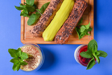 Different coloured eclairs on a wooden board over blue background, tiramisu and panna cotta dessert.