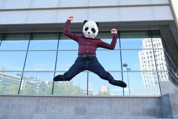 Young business man wearing a panda head mask, jumping in front of the a corporate building. Bizarre businessman.