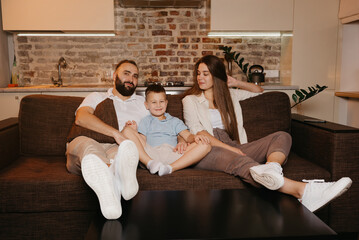 Dad, son, and mom are watching TV on the sofa in the apartment. The family is enjoying a happy evening at home. A young mother with long hair is smiling at her relatives.