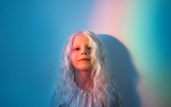 Child dream. Imaginary world. Bedtime fairytale magic. Portrait of happy cute blonde small girl with curly hair in rainbow color neon light on blue empty space background.