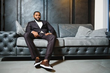 Young black man in suit on dark background sitting on a sofa. Fashion portrait of young man.