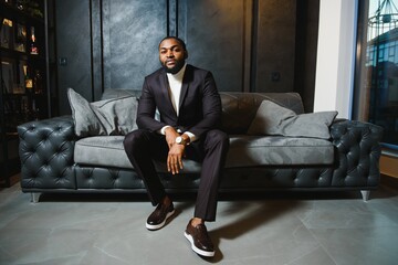 Young black man in suit on dark background sitting on a sofa. Fashion portrait of young man.