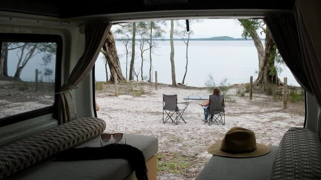 View out the back door of a campervan of a young couple enjoying coffee at a lake. 