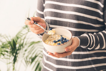 Woman eating tasty oatmeal with blueberry in room, closeup