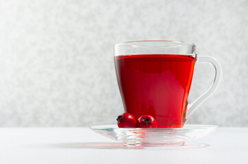 Red herbal tea in transparent cup with hawthorn berry in morning sunlight on soft light white wood table, copy space, closeup.