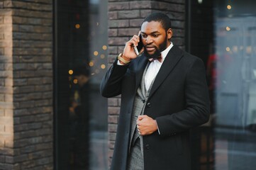 Business communications. Happy young African man in formal wear talking on the mobile phone and smiling while standing outdoors
