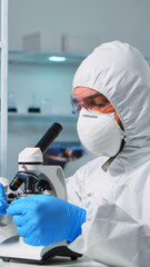 Scientist in ppe suit placing a slide on specimen stage of a laboratory microscope making adjustments. Chemist in coverall working with various bacteria, tissue blood samples for antibiotics research