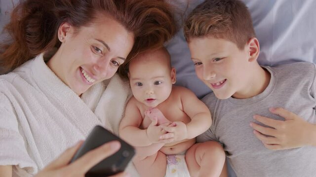 Happy mother with her kids are making a selfie or video call to father or relatives in a bed. Concept of technology, new generation,family, connection, parenthood, authenticity.