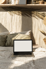 Blank screen laptop. Warm sunlight shadows on the wall. Parisian vibes. Warm bohemian style home interior design. Copy space mockup template. Hygge bedroom with bed, pillows, linens, decorations.