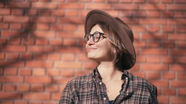 Slow-motion footage. Close-up portrait of an attractive cheerful woman in a hat and glasses looking to the camera.