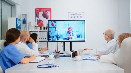 Team of doctors having video conference sitting at desk in hospital meeting room discussing health issues with remote specialist surgeon and taking notes. Nurse analysing diagnosis of patients