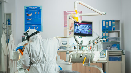 Dentist with face shield working in dental unit during covid-19 pandemic using sterilized tools. Orthodontist and nurse working wearing coverall, face shield, mask, gloves in stomatological office