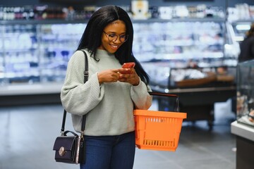 African american woman with shopping cart trolley in the supermarket store look on mobile phone.